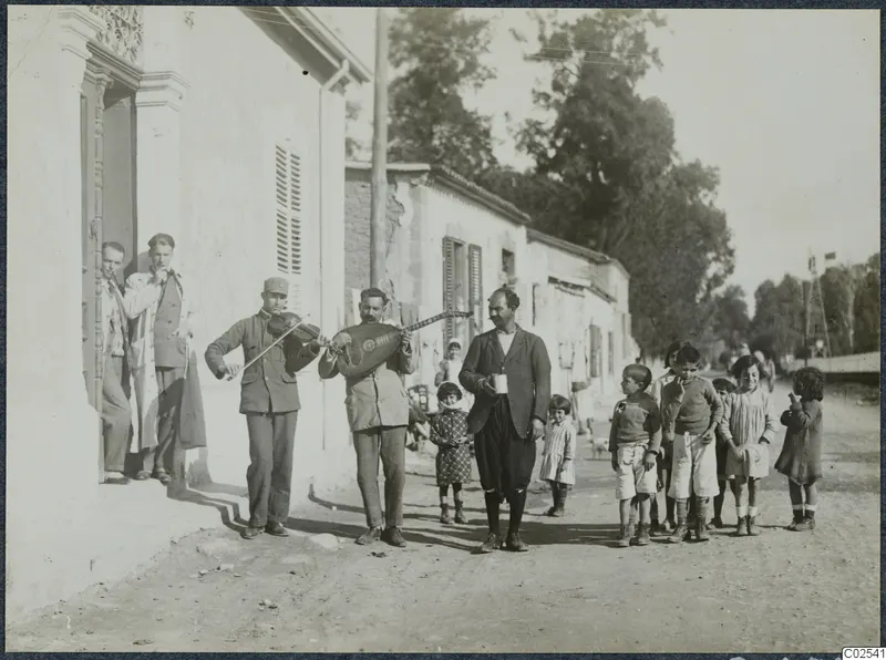 Two men play violin and lute in a street outside a whitewashed building, with children gathered around and a man holding a cup.