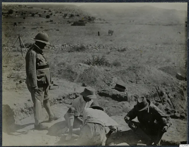 Men digging in a trench at Ayios Iakovos while a man in a pith helmet stands at the edge watching