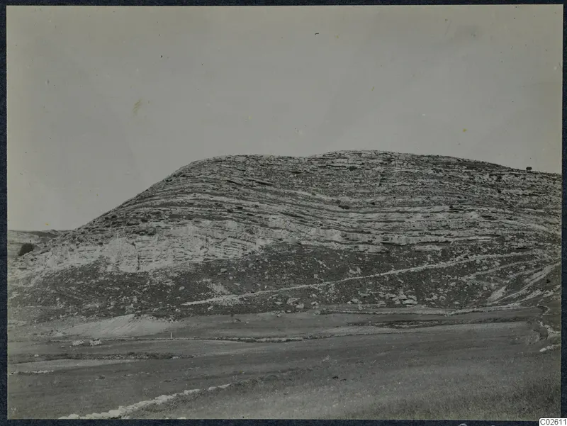 Layered limestone hill rising above open ground, with a faint track running across its slope.