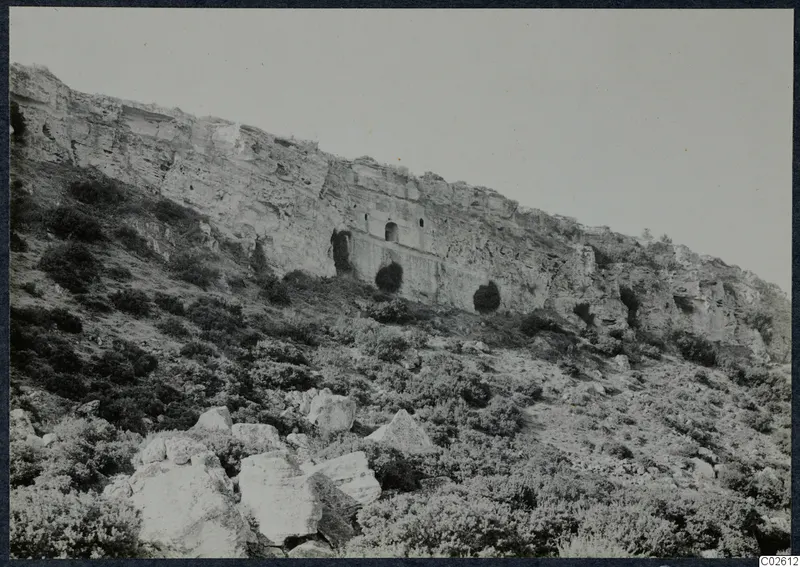 Pale limestone cliff with cut openings above a slope of bushes and boulders, Karpas peninsula