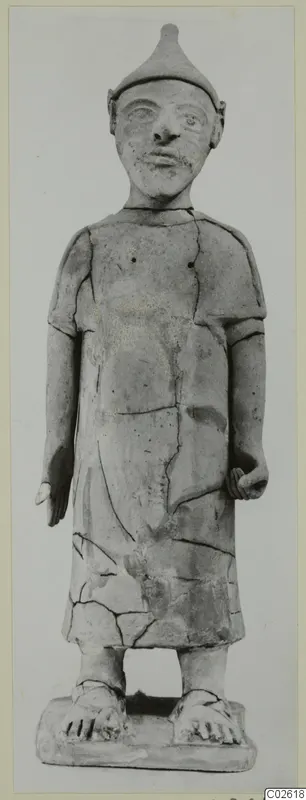 Terracotta standing figure in a pointed cap and short-sleeved robe, seen from the front, with cracks and visible joins.