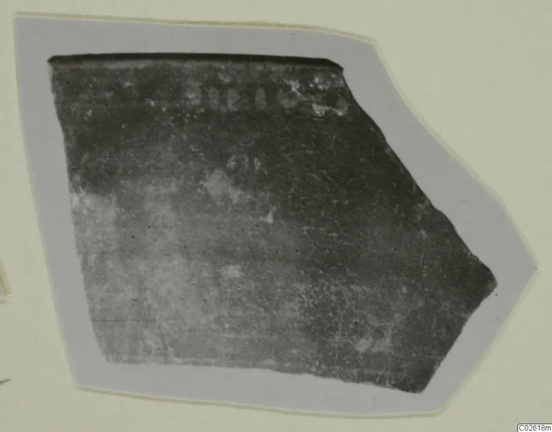 Fragment of a dark pottery sherd with a straight top edge and faint marks near the top.