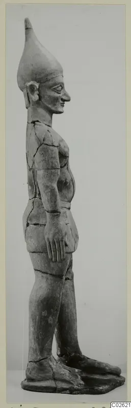 Standing terracotta figure in profile, with a tall conical headdress and arms held close to the body