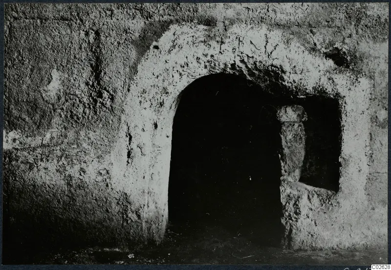 Arched opening of a built tomb chamber in a rough plastered wall, with a short upright stone post beside it