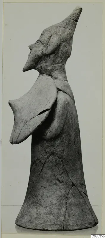 Terracotta figurine in profile, with a tall flaring body, a pointed head with a beaked face, and a flat projection at the shoulder.