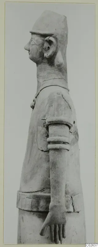 Profile photograph of a terracotta figure of a standing human figure with a tall cap, facing left, one arm bent.