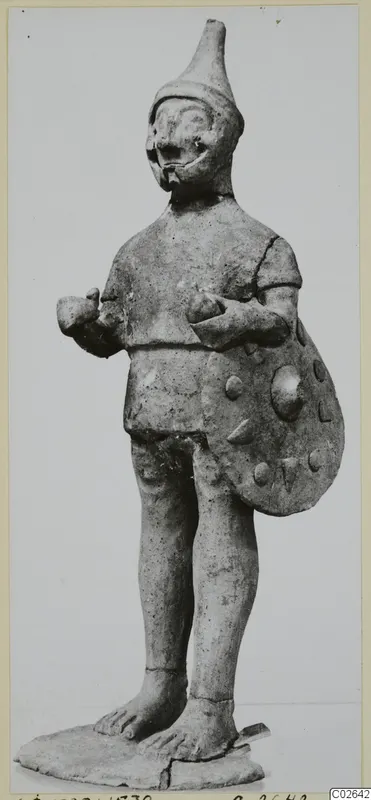 Terracotta figure of a standing figure with a tall pointed cap, holding a round shield with raised knobs, on a flat base