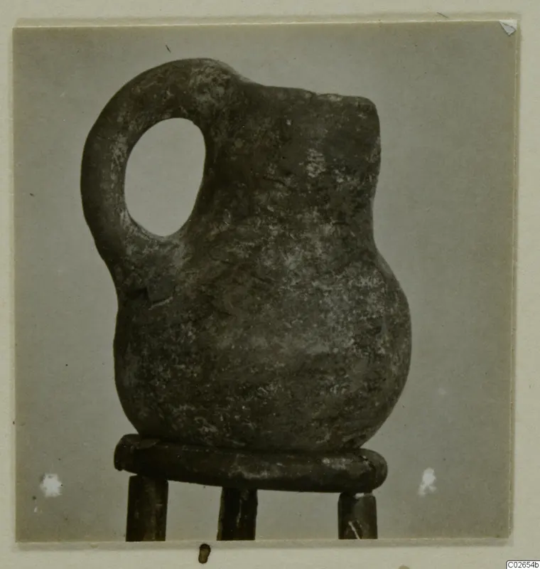Red Polished IV jug with a loop handle, standing on a wooden stand against a grey backdrop, from Ayios Iakovos