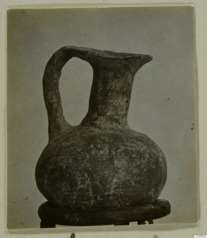 Jug with a loop handle, a flared spout-like rim and a rounded body, standing on a dark round stand against a grey backdrop.