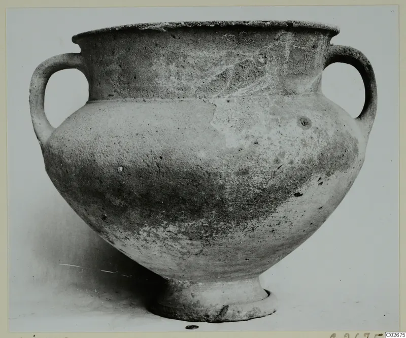 Two-handled wheel-made pottery jar with a wide body and short foot, photographed on a plain background.