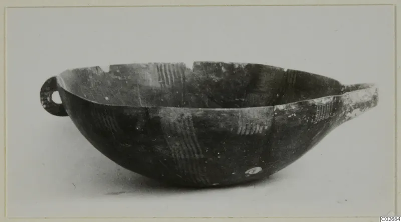 Dark pottery bowl with a small loop handle at left and light hatched bands in its decoration, photographed on a pale background.