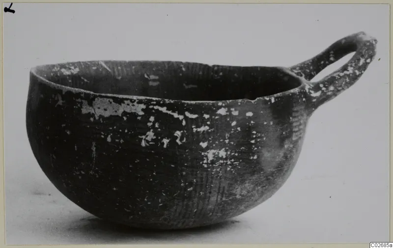 Dark pottery bowl with a single loop handle on its rim, shown in side profile against a pale background.