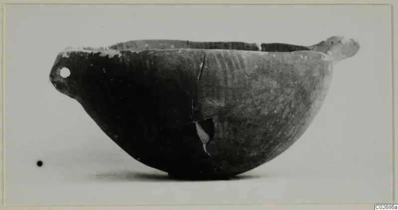 Side view of a dark pottery bowl with a pierced lug at the left and a horizontal handle at the right