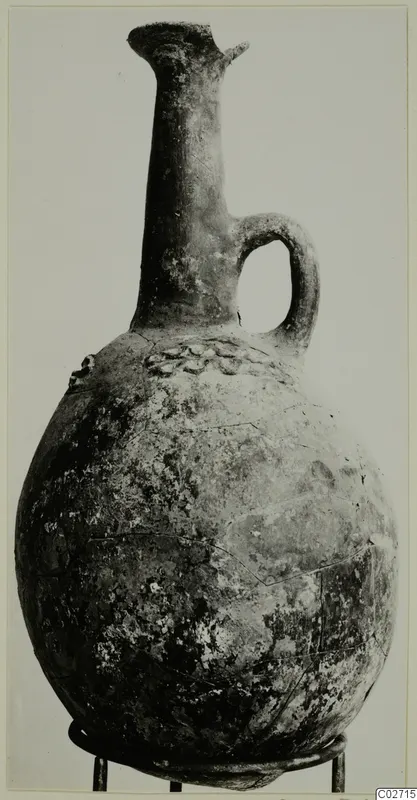 Red-polished jug with a tall narrow neck, a single loop handle and a rounded body, photographed on a stand.