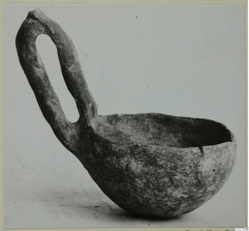 Red polished pottery bowl with a tall loop handle, photographed against a plain grey background.