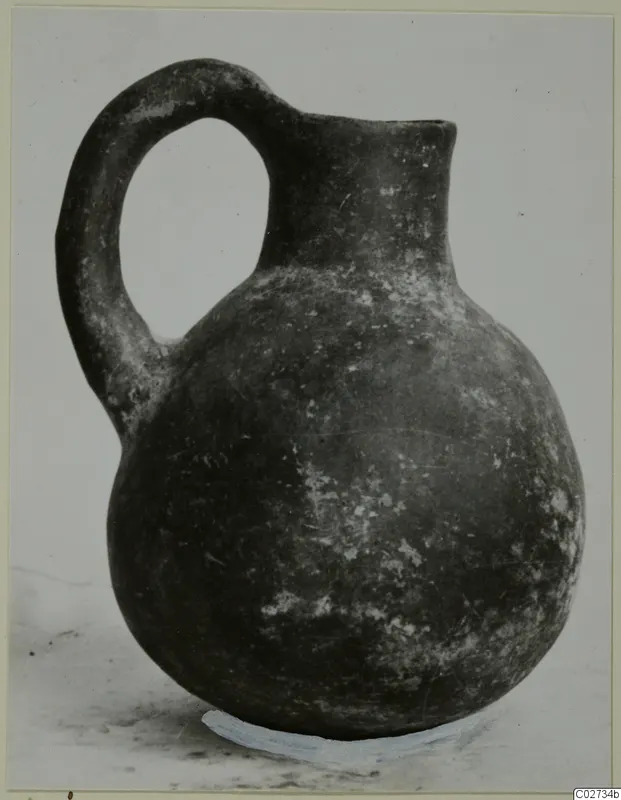 Round-bodied jug with a high loop handle and short neck, dark surface with pale flecks, photographed against a grey backdrop.