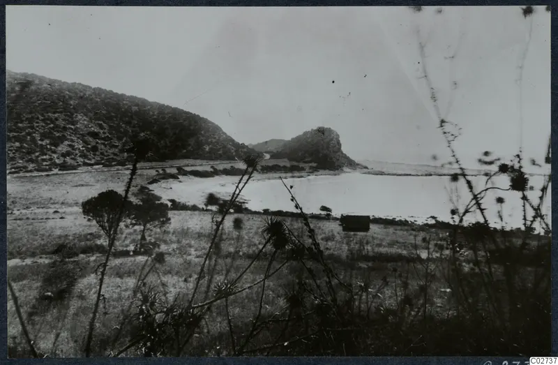 Landscape at Vouni with a scrub-covered hillside, a rocky knoll and a pale expanse of water behind grass in the foreground.