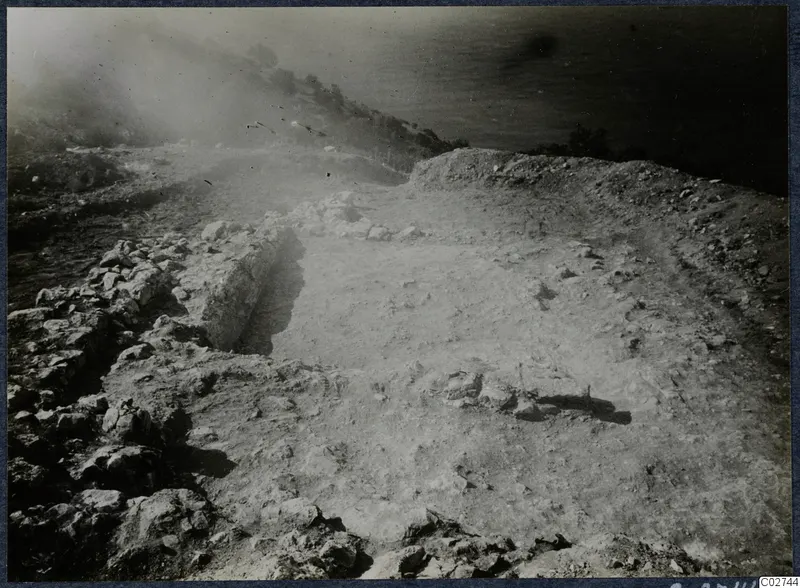 Excavated clifftop at Vouni with stone wall foundations, a cleared surface, and the sea and scrubby slope beyond