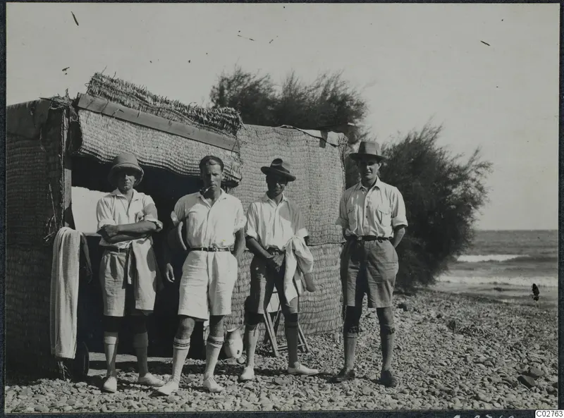 Four men in shorts and light shirts stand before a reed-mat hut on a pebble beach by the sea.