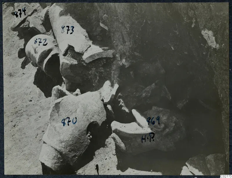 Broken carved stone fragments, probably sculpture parts, lying in an excavated area with numbered labels