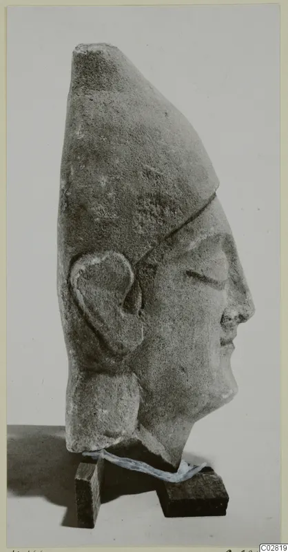 Profile view of a limestone head with a tall headdress and carved ear, from Kition, set on two small wooden blocks.
