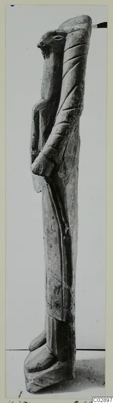 Limestone sculpture fragment of a tall standing figure with a beak-like head, photographed on a mounted card