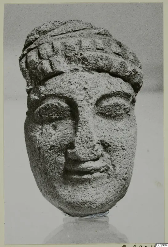 Limestone head of a figure with a banded headdress, nose and mouth worn, shown against a grey backdrop.