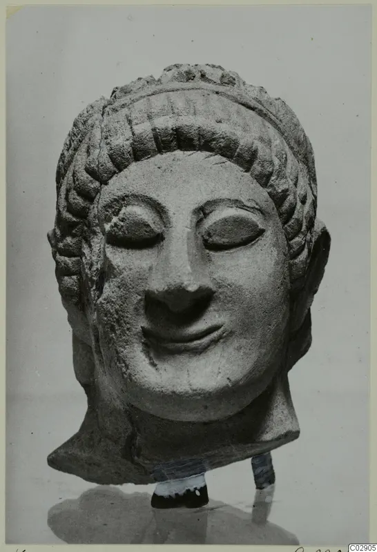 Limestone head of a figure with a tiered hairstyle and closed eyes, shown on a stand against a plain grey backdrop.