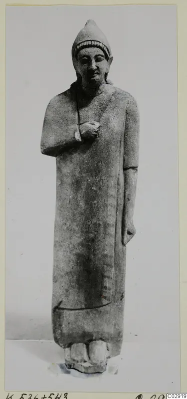 Limestone statue of a standing figure from Kition, with a tall cap and arms folded across the chest.