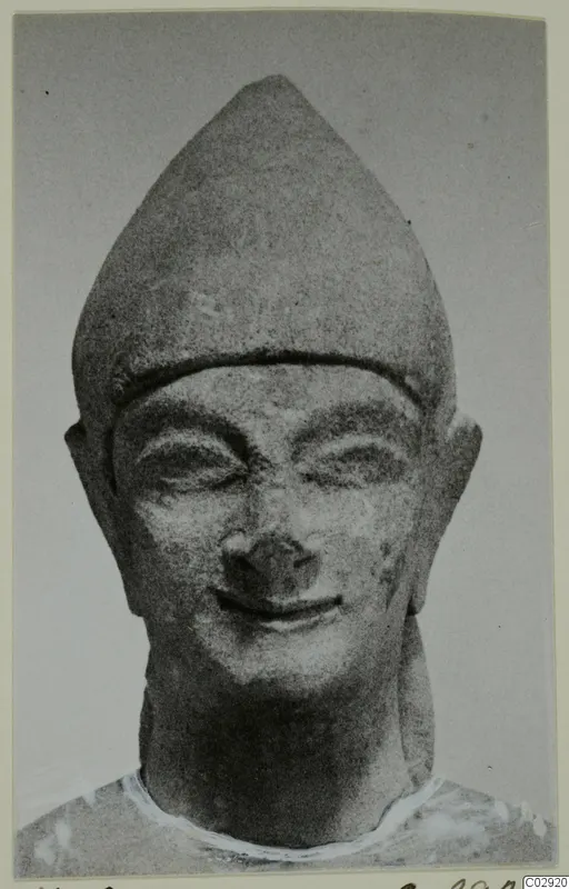 Limestone head of a statue from Kition, wearing a tall pointed cap, with a broad smile and narrowed eyes
