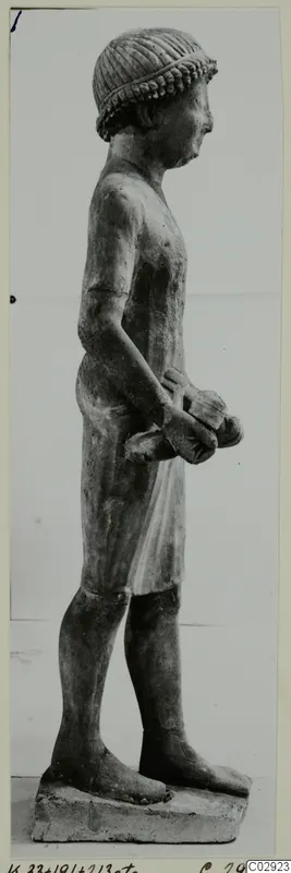 Limestone statue of a standing male figure in profile, wearing a cap and a short kilt, holding an object in his hands.
