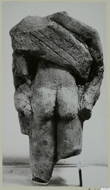Back view of a limestone statue of a standing figure with arms at its sides and a block-like upper part.