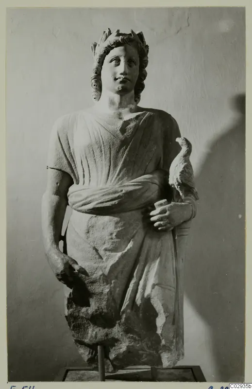 Carved stone statue of a standing figure with a leafy wreath, holding a small bird against its side, on a square base.