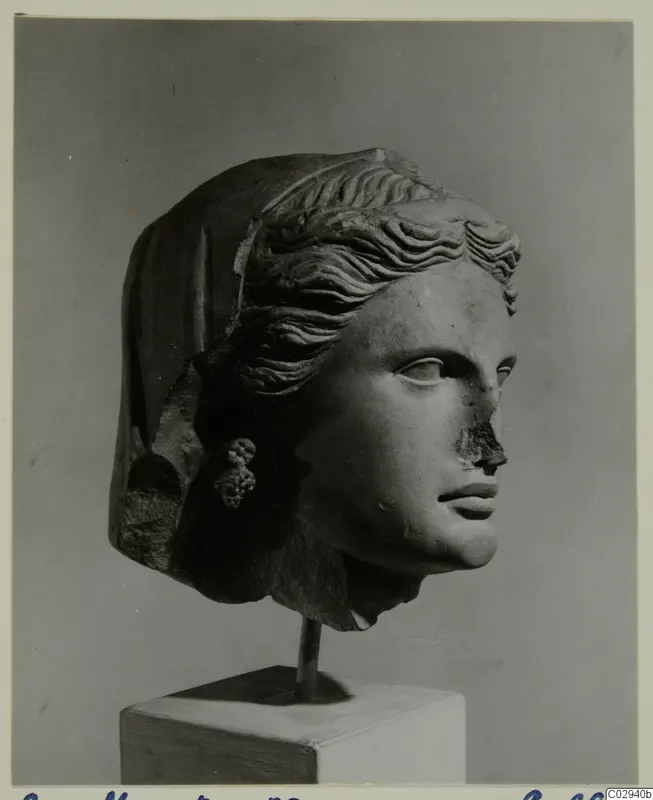 Stone female head with a veil, wavy hair and a cluster-shaped earring, mounted on a rod above a white plinth against grey