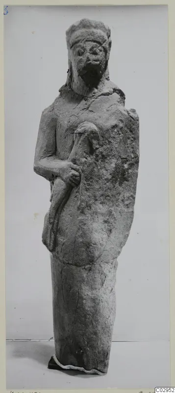 Limestone statuette of a standing bearded figure with a wide headdress, one hand holding a curved object against the body.