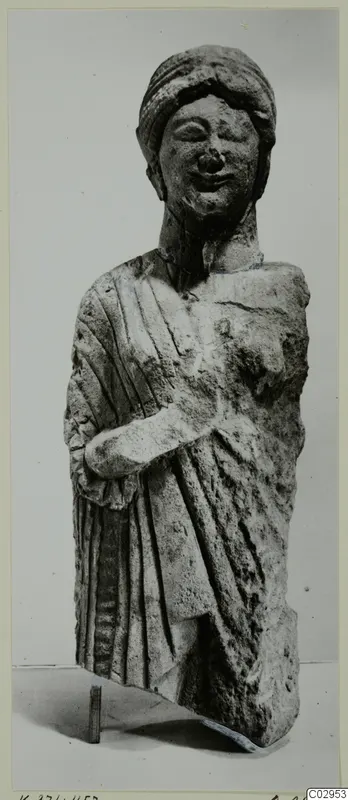 Limestone statue of a draped standing figure with a head covering, right arm bent across the body, lower part broken away