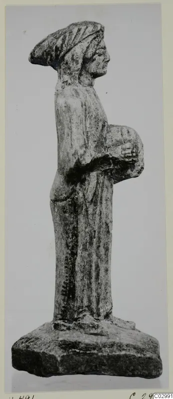 Limestone figurine of a standing woman holding a round object at her side, on a rectangular base, seen in profile.