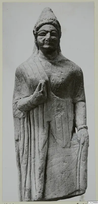 Standing limestone statue of a figure in a long robe with a conical headdress and one hand raised to the chest