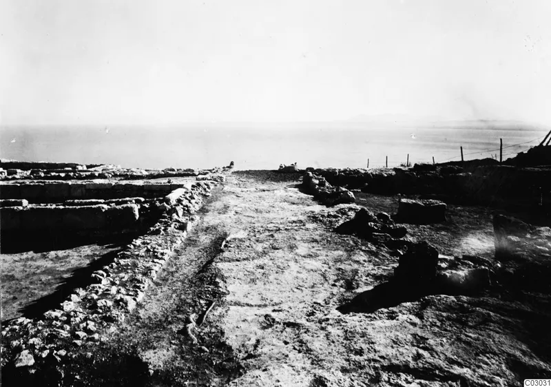 Stone wall foundations and a dirt path running toward the sea at Vouni, with a wire fence on the right.