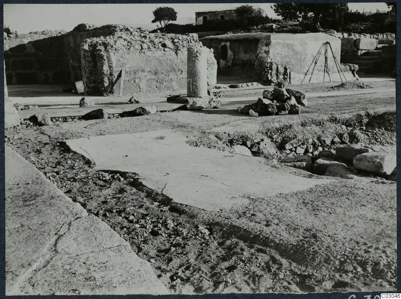 Ruined walls, a column drum and a heap of stones at the palace at Vouni, with trees and a house behind.