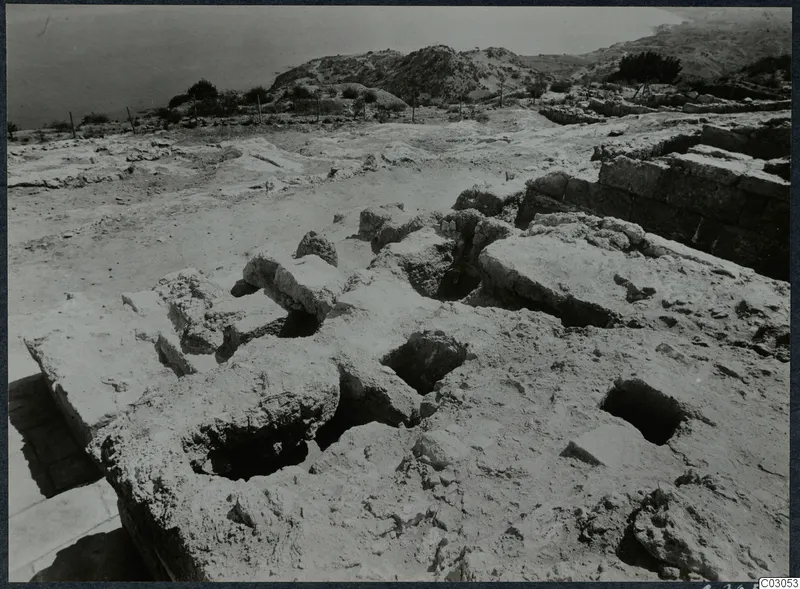 Excavated stone walls and rectangular pits at Vouni, with a row of stakes along the edge and the sea and hills beyond.