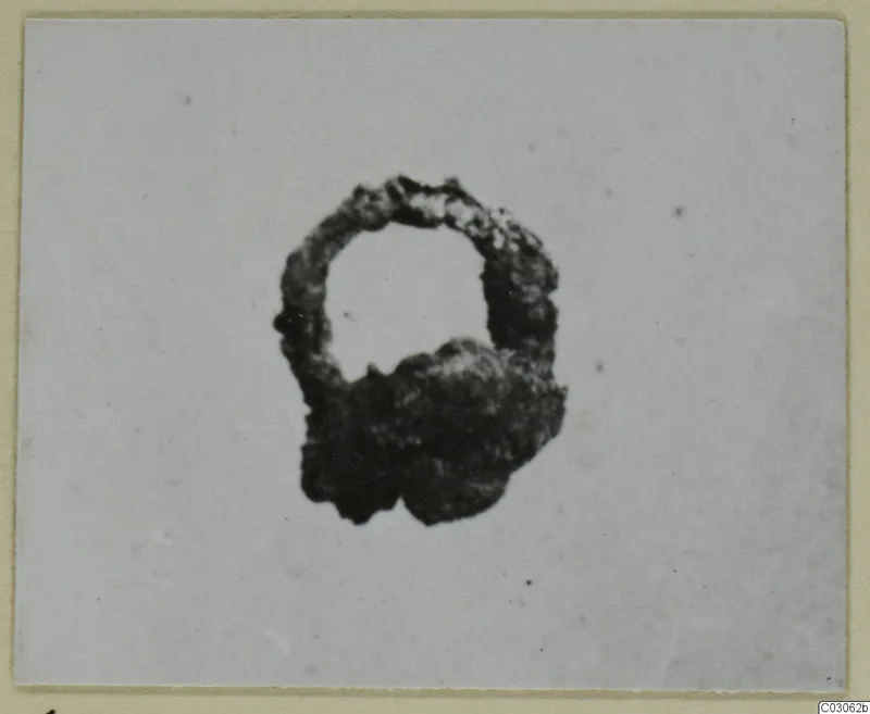 Heavily corroded iron object with an open loop at the top and a lumpy mass at its base, photographed on a card mount