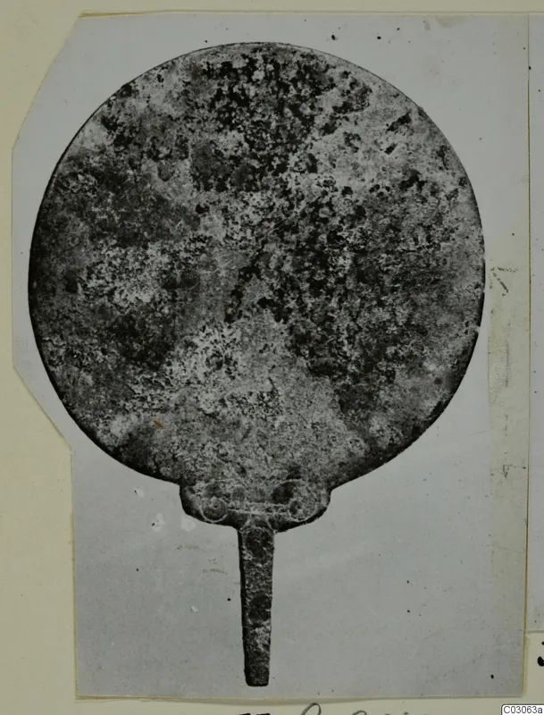 Corroded round metal disc with a narrow tang, photographed on a grey card mount, from Marion Tomb 41