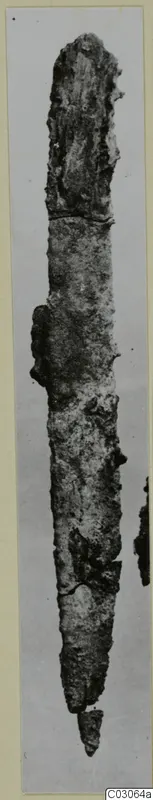 Long corroded iron knife, photographed upright, with a small fragment broken off near its tip.