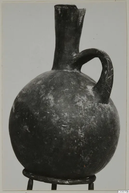 Dark round-bodied jug with a tall neck, a single loop handle and a pouring lip, standing on a metal stand.