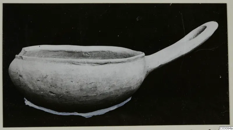Plain white hand-made pottery bowl with a long handle, photographed against a dark background.
