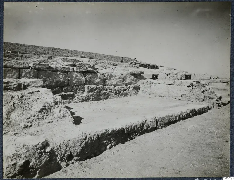 Stone wall foundations and a flat floor area at Vouni Palace, with two small figures standing on the ridge behind