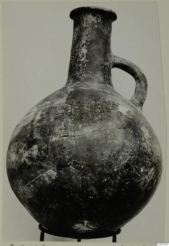 Pottery Red Polished III jug with a single loop handle, standing on a metal stand against a plain background