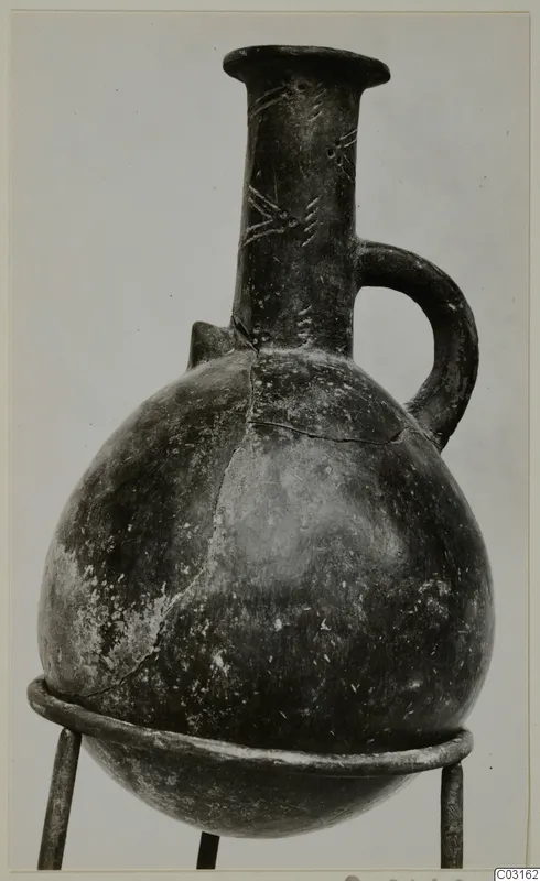 Red Polished III jug from Lapithos, with a single handle and incised neck decoration, shown on a metal stand