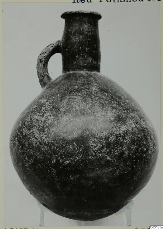 Red Polished III jug with a single loop handle and rounded body, shown on a small stand against a plain grey backdrop